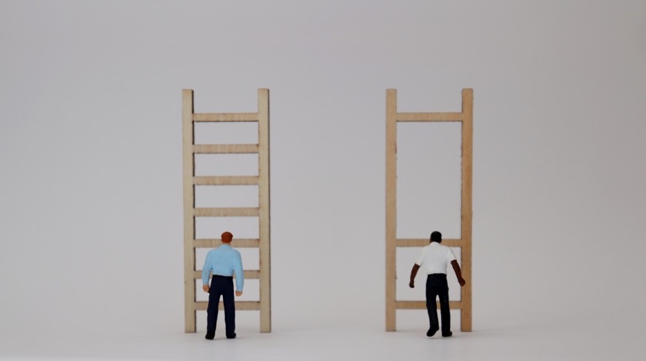 Picture of two people standing in front of ladders. The white person has a ladder full of rungs. The Black person's ladder only has three rungs, making it extremely difficult, if not impossible to reach the top. 
